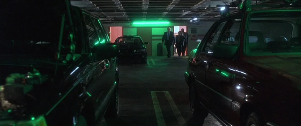 The Usual Suspects movie still: wide — A low-lit underground parking garage features rows of cars and concrete pillars. Three men in suits walk…