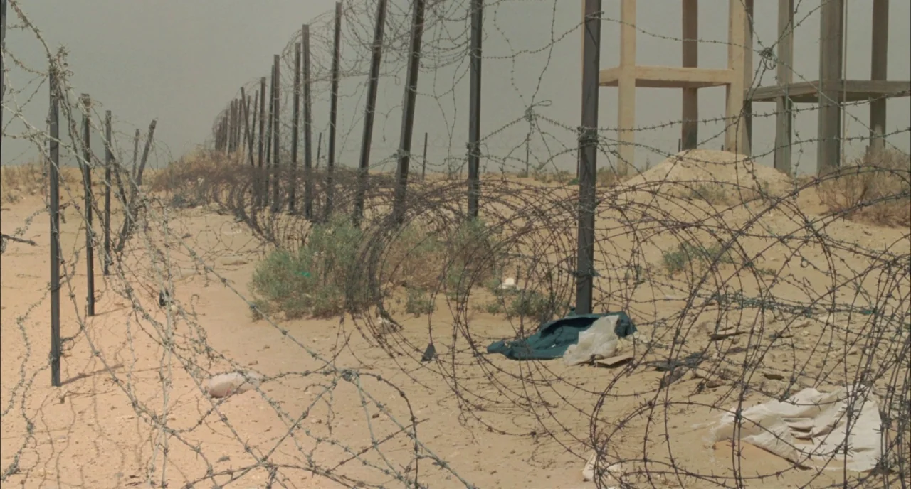 Incendies movie still: wide — A vast sandy desert features a long line of barbed wire fencing. A skeletal concrete water tower structure stands near…