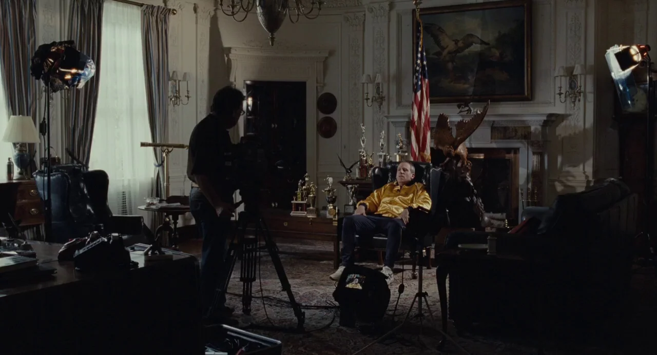 Foxcatcher (2014) movie still: wide — A room with wooden furniture and ornate wall molding featuring an American flag. A man stands at a video…