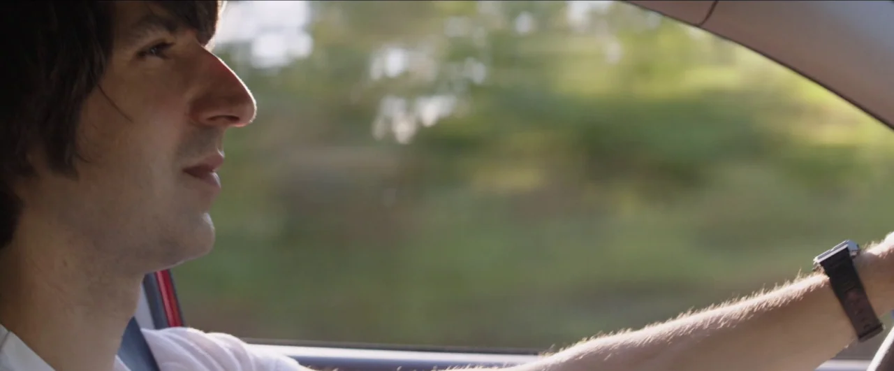 Dean (2016) movie still: closeup — A view inside a car moving through a rural or suburban setting. A man with dark hair drives while looking out the…
