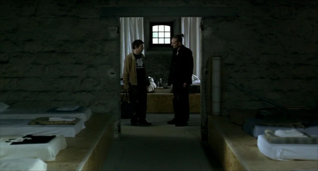 On War (2008) movie still: wide — A stone-walled room features multiple low wooden platforms holding simple white mattresses. Two men stand in the…
