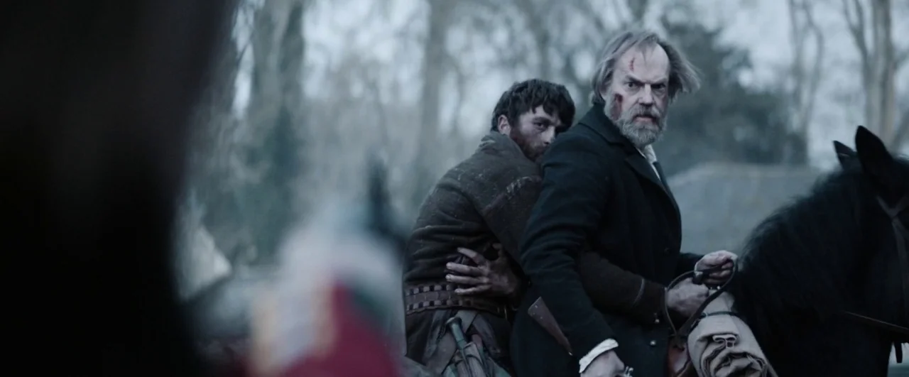Black ’47 (2018) movie still: medium — A desolate wooded area during an overcast day provides a bleak backdrop. Two men on horseback are fleeing…
