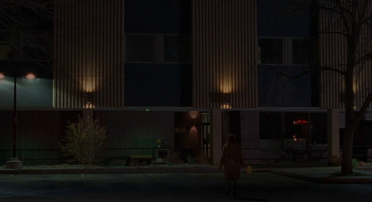 Certain Women (2016) movie still: wide — An empty parking lot at night sits in front of a multi-story office building. A woman walks alone toward…