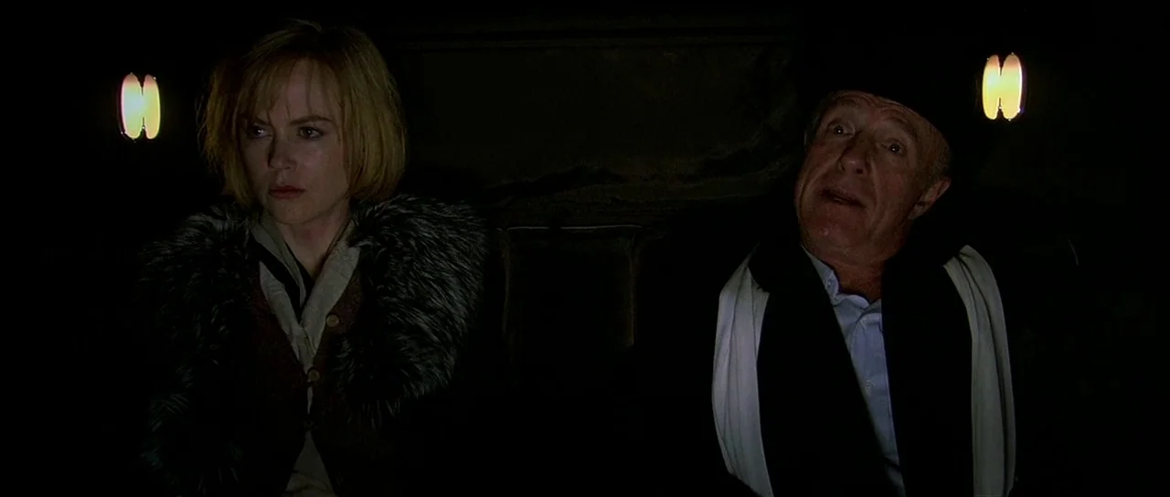 Dogville (2003) movie still: medium — A dark, cramped interior of a moving vehicle during the night. A blonde woman and an older man are seated next…