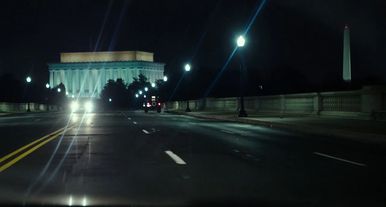 Last Flag Flying (2017) movie still: wide — A wet asphalt road at night with distant illuminated monuments under dark skies. A single vehicle moves…