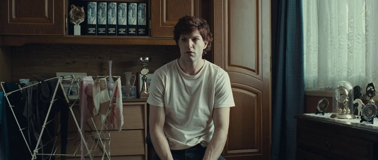 Our Day Will Come (2010) movie still: medium — A furnished space with wood cabinets, a laundry drying rack, and a bedside table. A young man sits in…