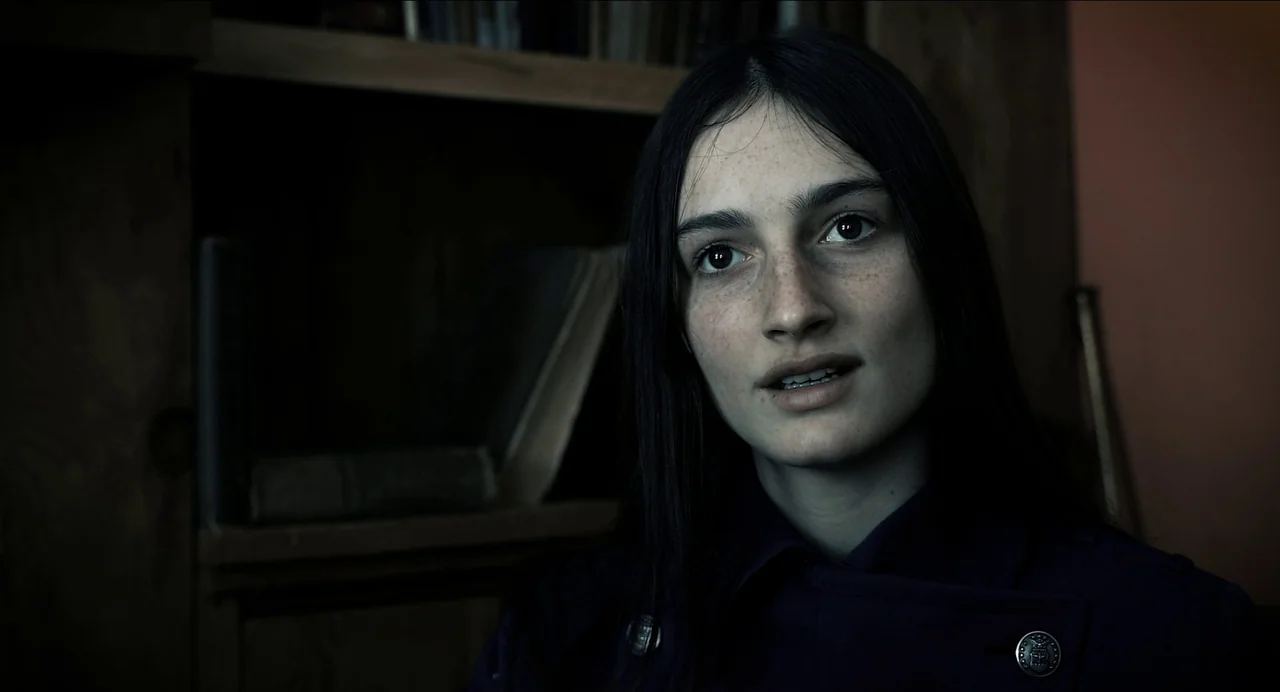 Where The Devil Roams (2023) movie still: closeup — A dark, dimly lit study features a wooden bookshelf with few items in back. A woman with long…