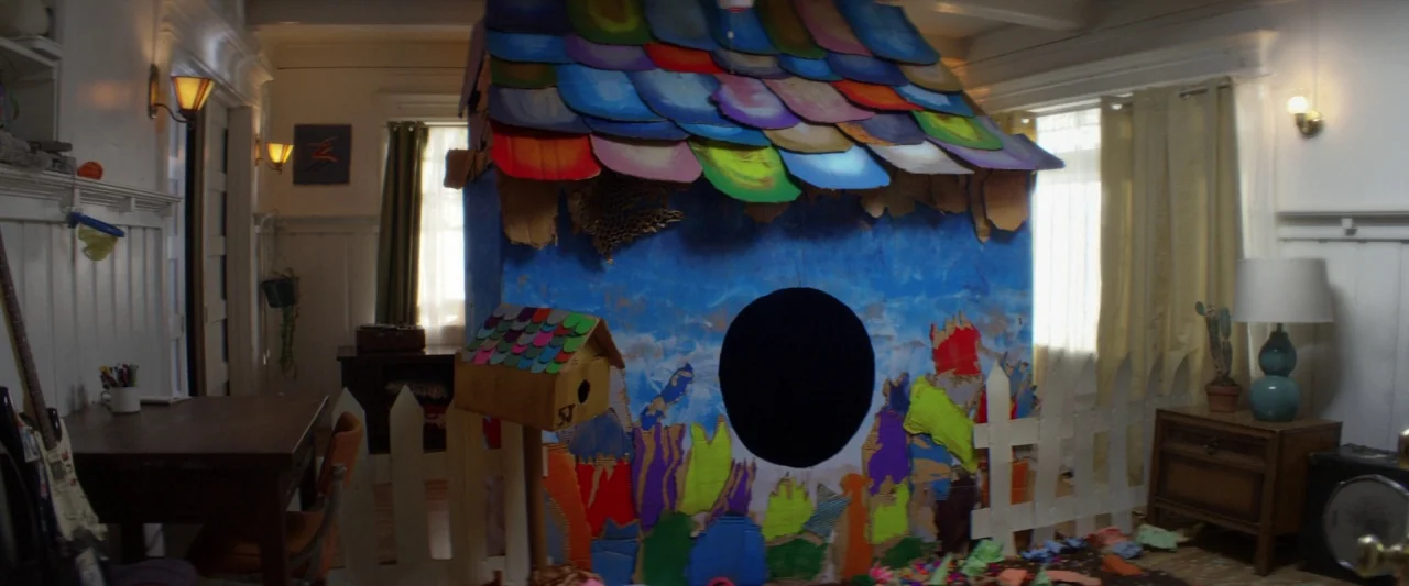 Seven Stages to Achieve Eternal Bliss (2018) movie still: wide — A homemade cardboard structure with a colorful shingled roof stands in a room. The…