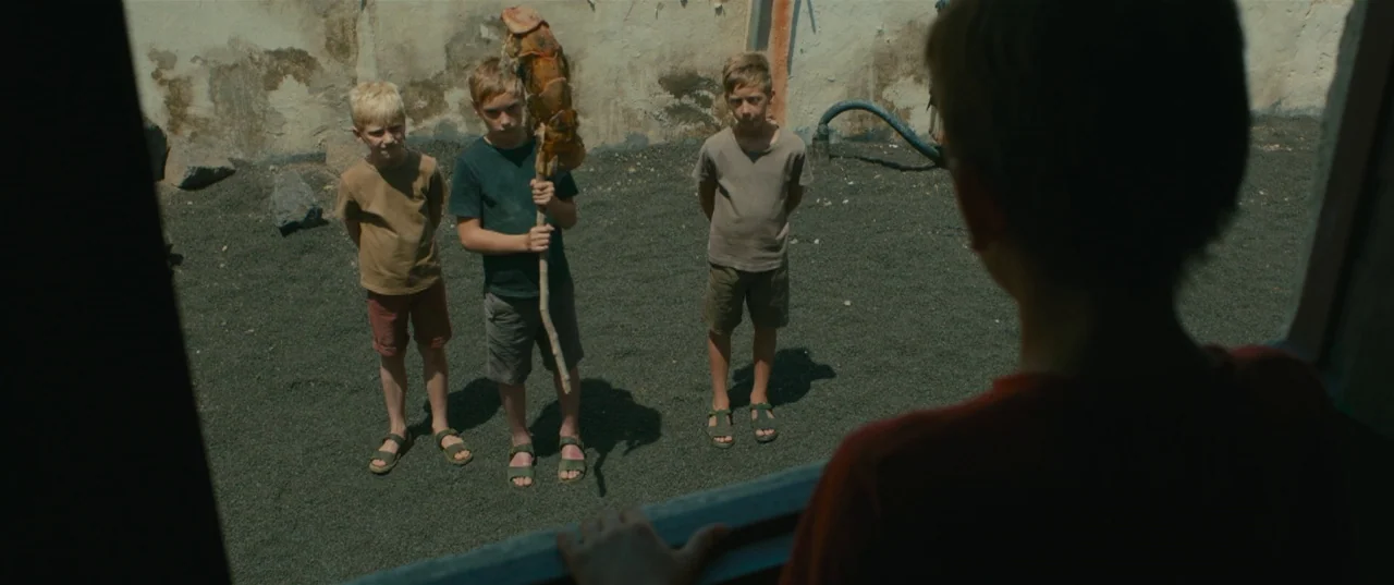 Evolution (2015) movie still: medium — A stark courtyard with high, weathered concrete walls surrounds a dark, gravel-covered ground area. Three…