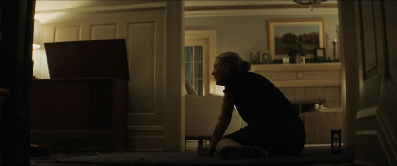 Vicious (2025) movie still: medium — A dimly lit residential living room features a fireplace mantel with framed artwork above. A woman kneels on…