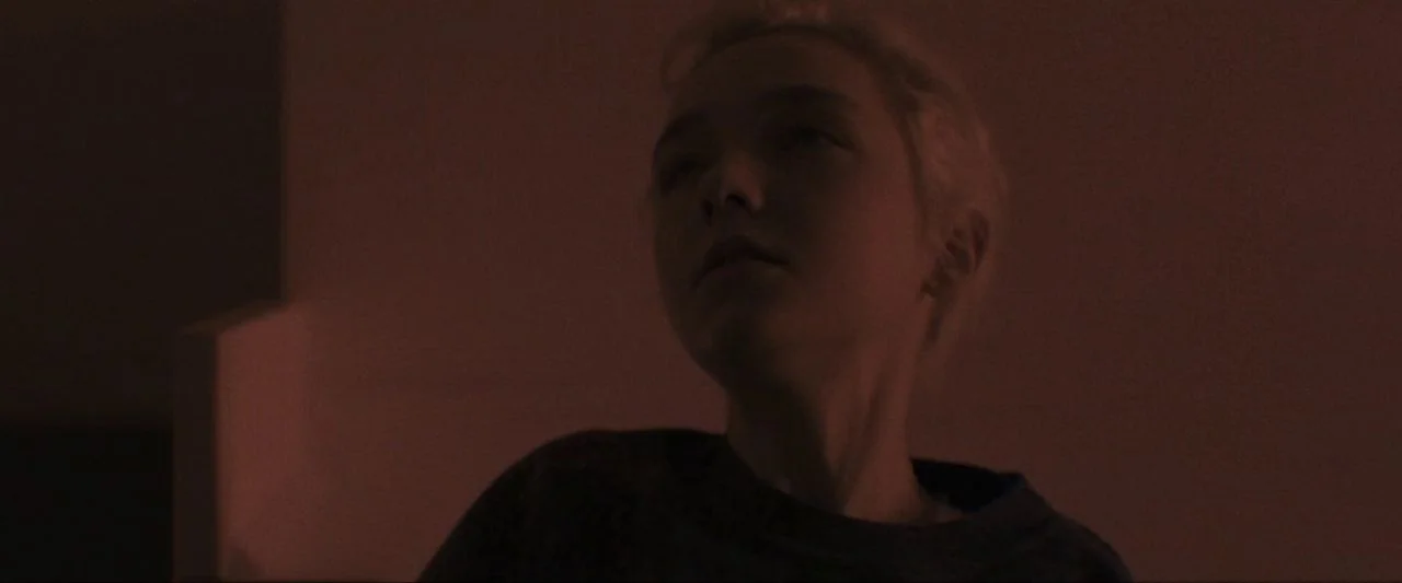 Come True (2020) movie still: closeup — A dimly lit room with walls cast in a warm, deep reddish tone. A young woman with short blonde hair tilts…