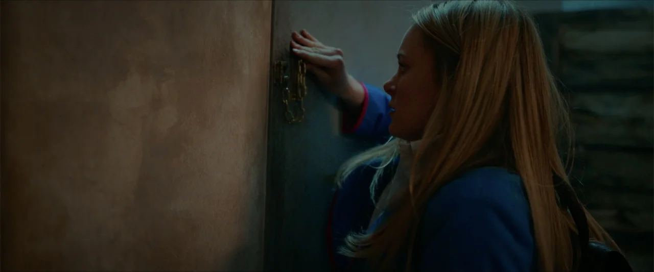 Love Me Love Me (2026) movie still: medium — A dark interior room features a plain wall and a wooden door with a chain lock. A young woman with…