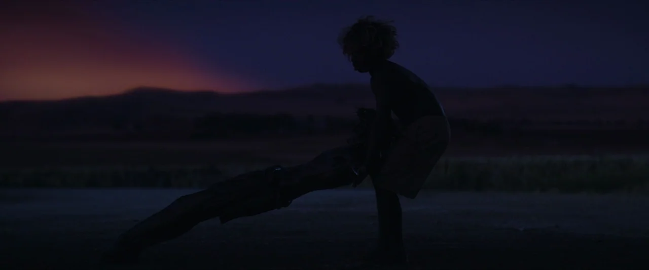 The New Boy (2023) movie still: fullbody — A vast, dark field at dusk with rolling hills visible against a twilight sky. Two teenagers stand in…