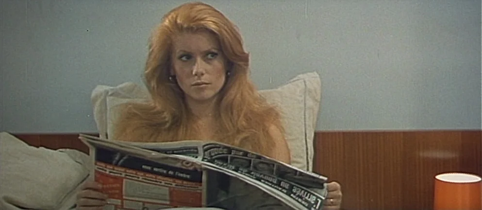 Mississippi Mermaid (1969) movie still: medium — A woman sits upright in a bed with a wooden headboard and light bedding. She holds a newspaper open…