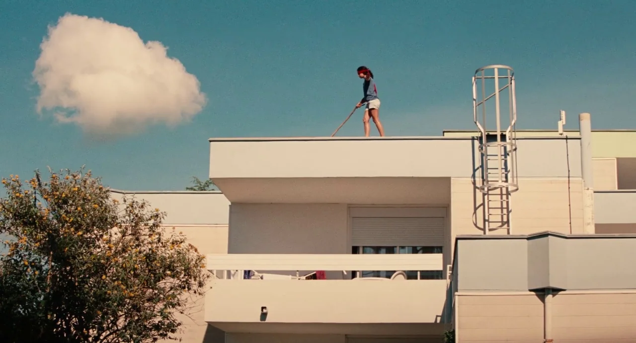 Ava (2017) movie still: wide — A modern flat-roofed residential building stands under a clear blue sky with one cloud. A young woman cleans the…