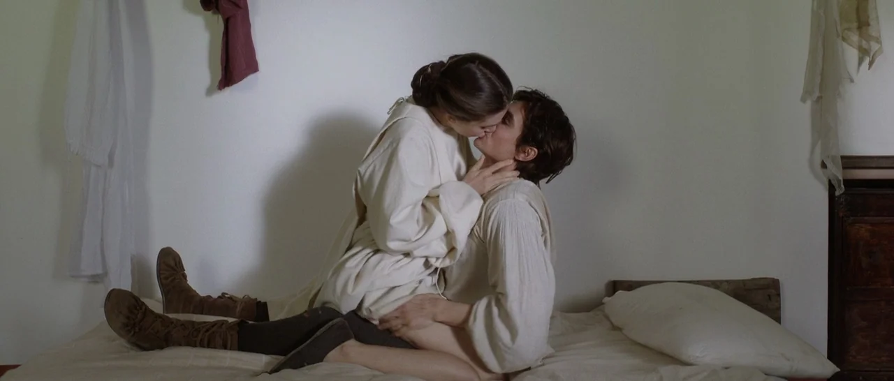 The Little Hours (2017) movie still: medium — A plain room with white walls and minimal decor contains a simple bed. A young man and a woman in…