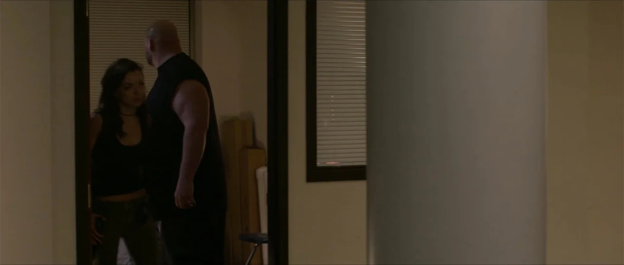 The Vault (2017) movie still: medium — A dimly lit room features a doorway leading into a space with closed window blinds. A large muscular man…