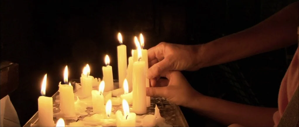 Genova (2008) movie still: closeup — A collection of white wax candles burns on a metal tray in a dark space. A person's hand places a new candle…