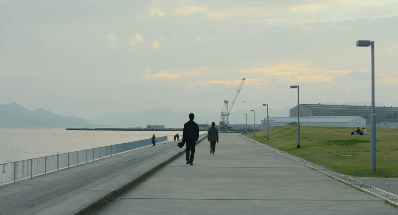 Drive My Car (2021) movie still: wide — A long concrete waterfront promenade stretches along the coast during a cloudy sunset. Two men walk away…