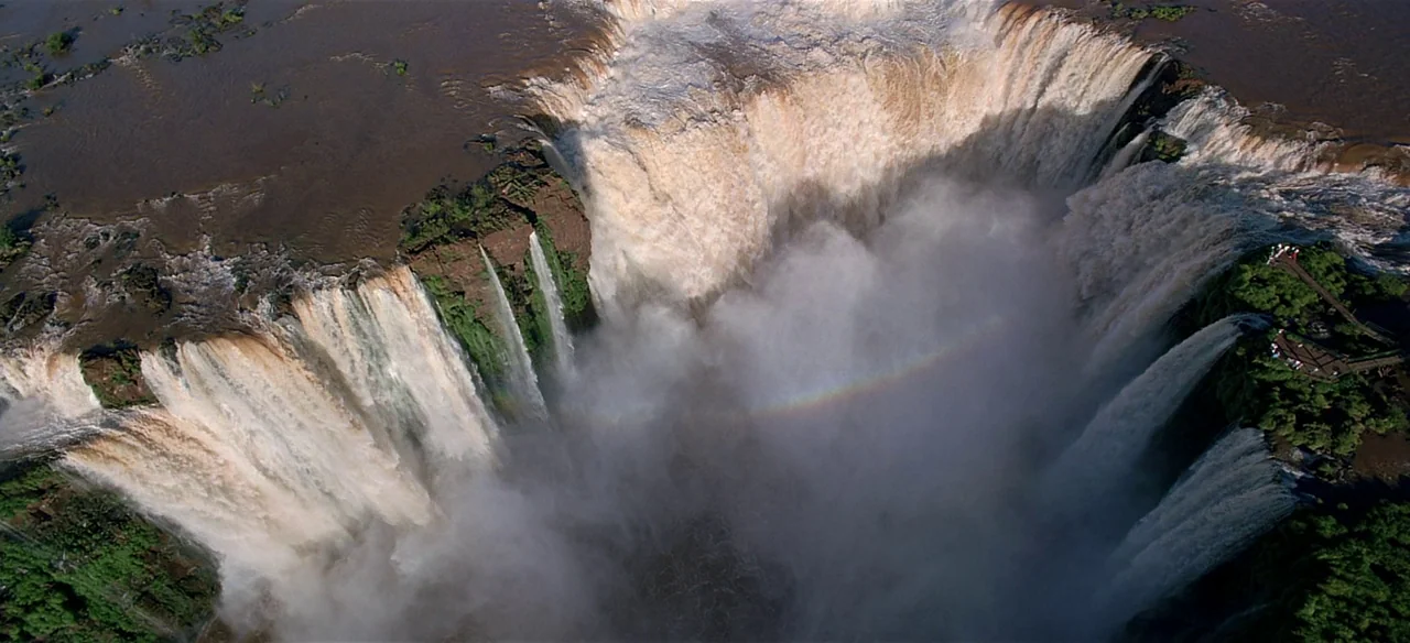 Baraka (1992) movie still: establishing — A wide aerial view shows a massive waterfall plunging into a deep basin. Mists rise from the bottom where…