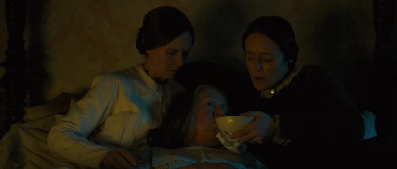 A Quiet Passion (2016) movie still: medium — A dimly lit room with dark wooden bedposts and patterned wallpaper. Two women hold a white cup for an…