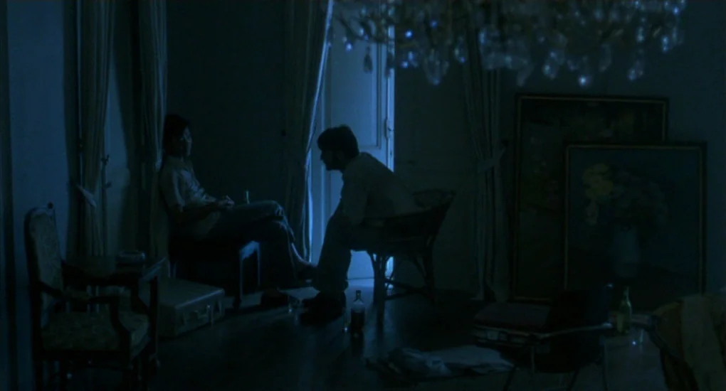The Killing Fields (1984) movie still: wide — A dimly lit hotel room with dark furniture and a partially visible crystal chandelier. Two men sit…