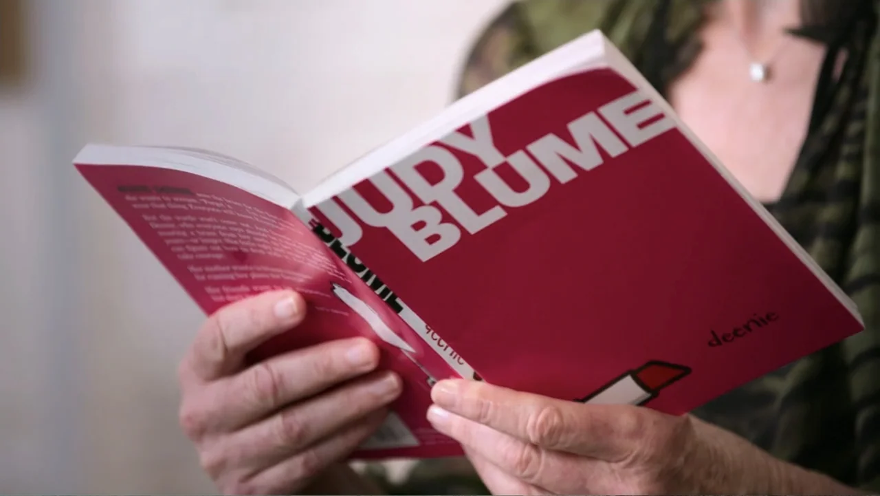 Judy Blume Forever (2023) movie still: closeup — A person holds a bright pink paperback book titled Judy Blume Deenie in their hands. The individual…
