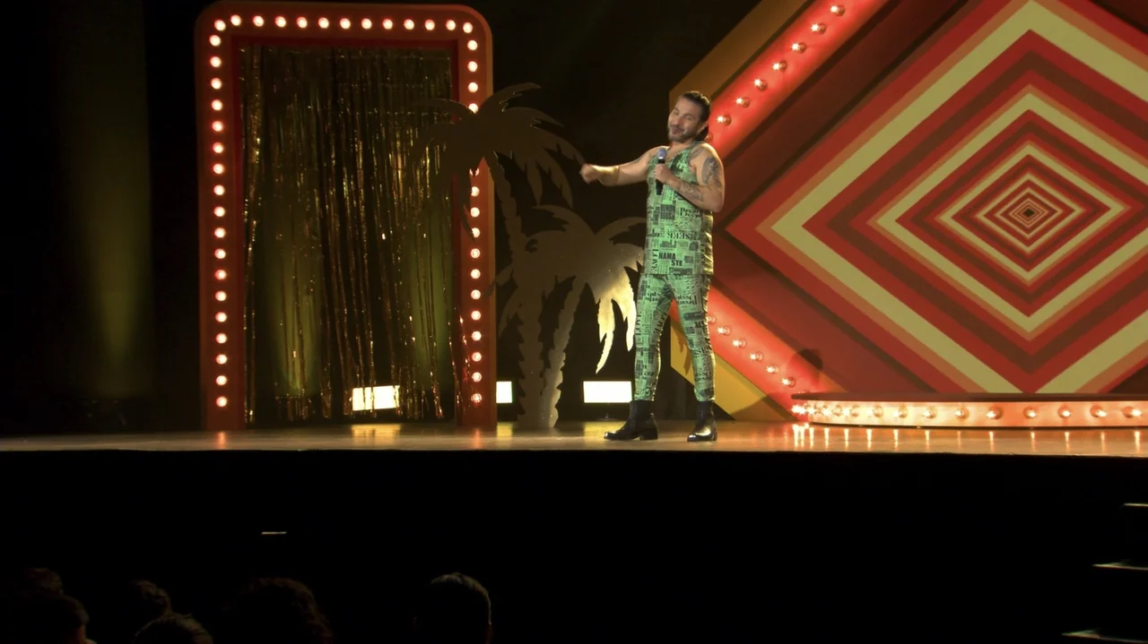 Felipe Avello: Doglover (2024) movie still: fullbody — A man stands on a bright stage decorated with palm tree cutouts and geometric light panels.…