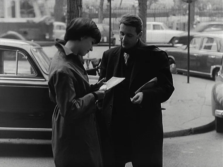 Vivre Sa Vie (1962) movie still: medium — A man and woman stand on a sidewalk surrounded by parked mid-century automobiles. They are holding papers…