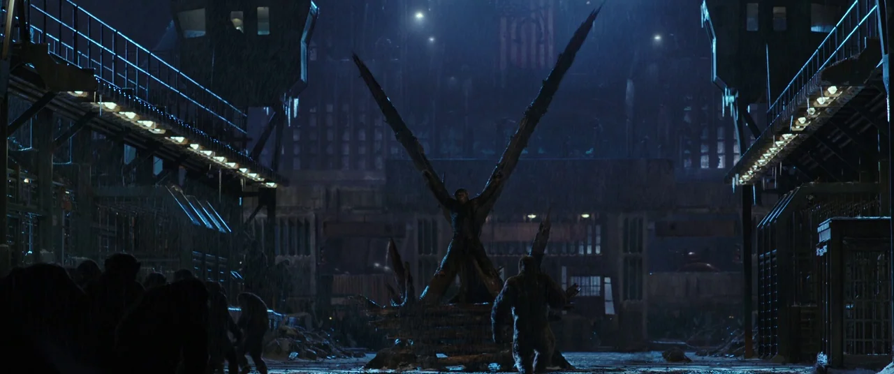 War for the Planet of the Apes (2017) movie still: wide — A dark, rainy industrial camp at night with metal structures and rows of apes. One ape is…
