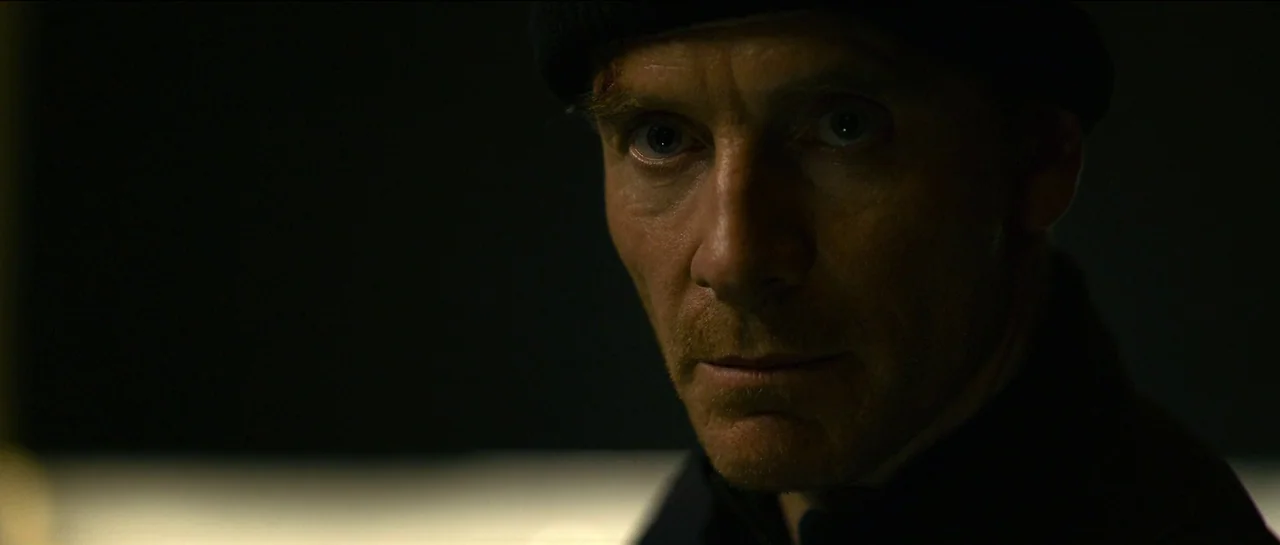 The Killer (2023) movie still: closeup — A dark, dimly lit environment features minimal light hitting a person's face. A man in a dark beanie stares…