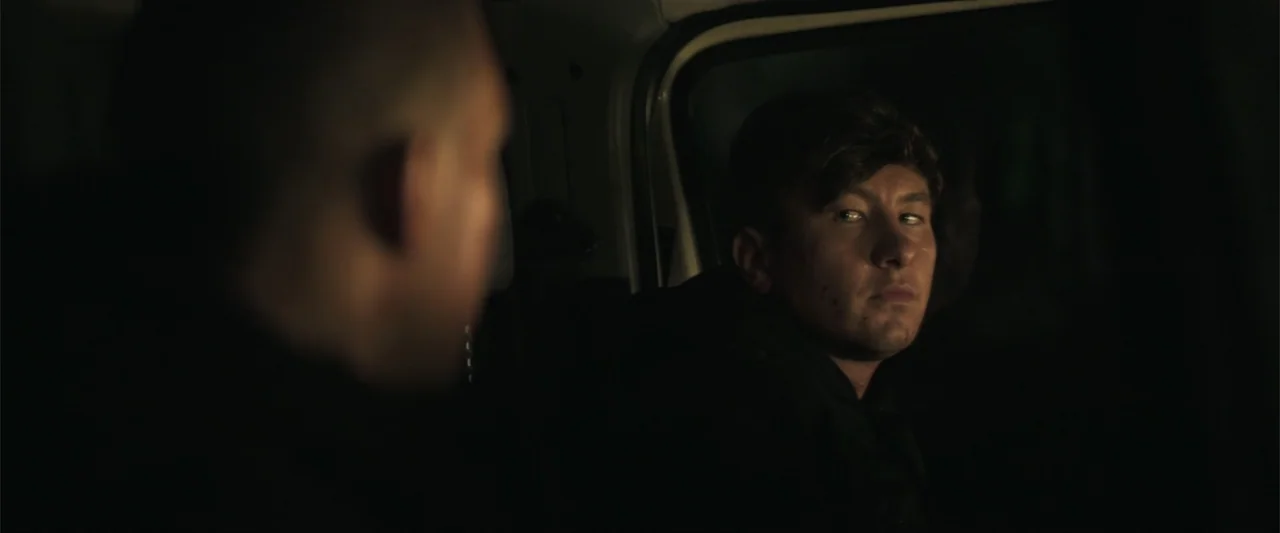 Bring Them Down (2025) movie still: medium — Two men sit inside the dark cabin of a moving van at night. The younger man in the passenger seat looks…
