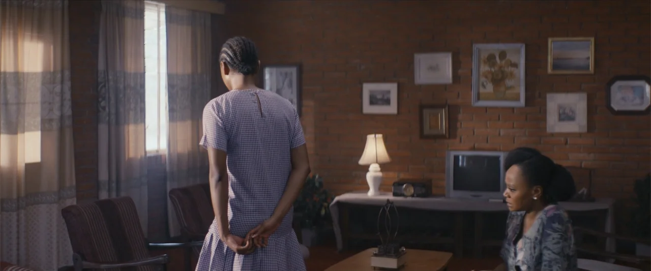 Ms. Kanyin (2025) movie still: medium — A living room with brick walls features numerous framed pictures and a television. A woman stands facing the…