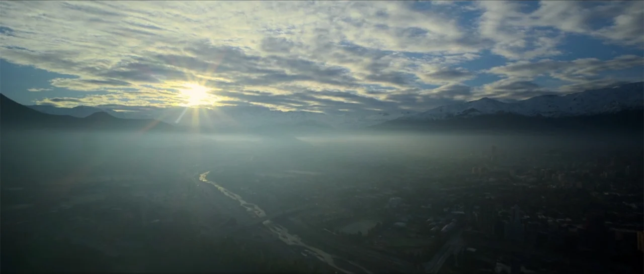 Disconnected 2 (2025) movie still: establishing — A wide aerial view of a city nestled in a misty valley below snowy mountains. The sun breaks over…