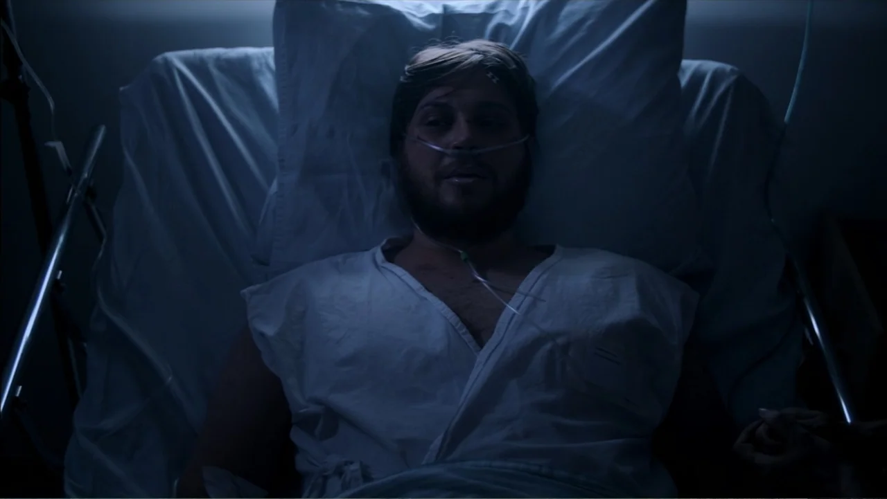 The Grotto (2022) movie still: medium — A dimly lit hospital room features a single bed with white linens and pillows. A man lies in the bed with a…