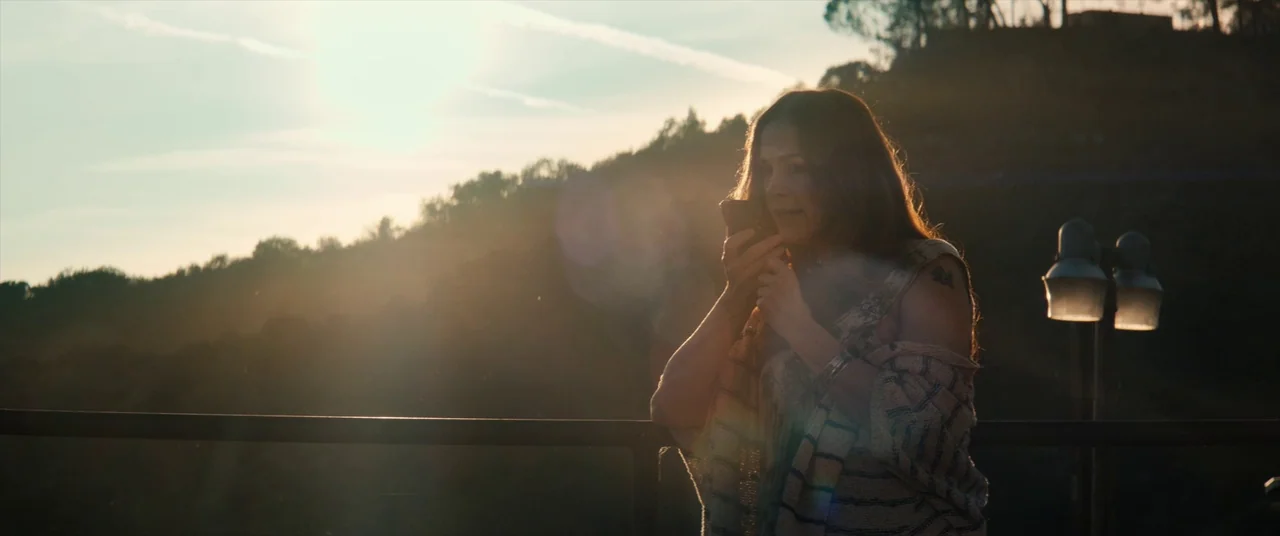 Tone Deaf (2019) movie still: medium — A woman stands on a balcony during golden hour overlooking a silhouetted hillside. She holds a smartphone to…