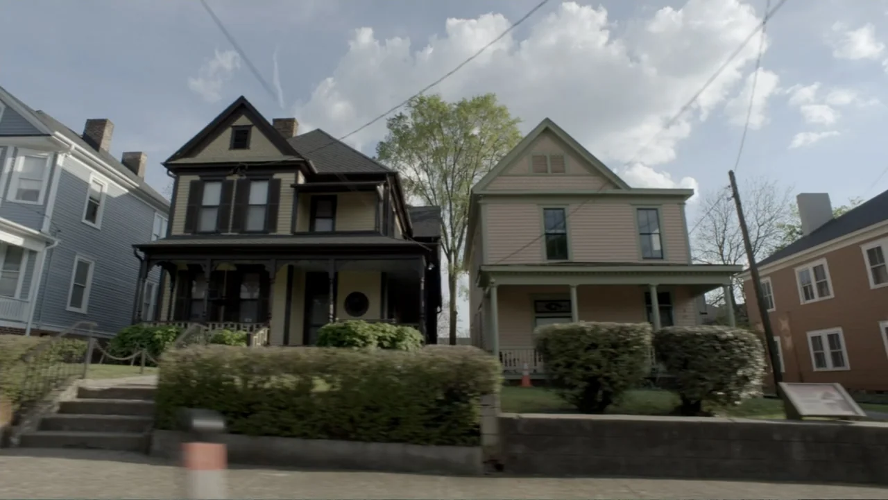 King in the Wilderness (2018) movie still: establishing — A row of historic houses stands along a sidewalk under a bright daytime sky. Thick green…