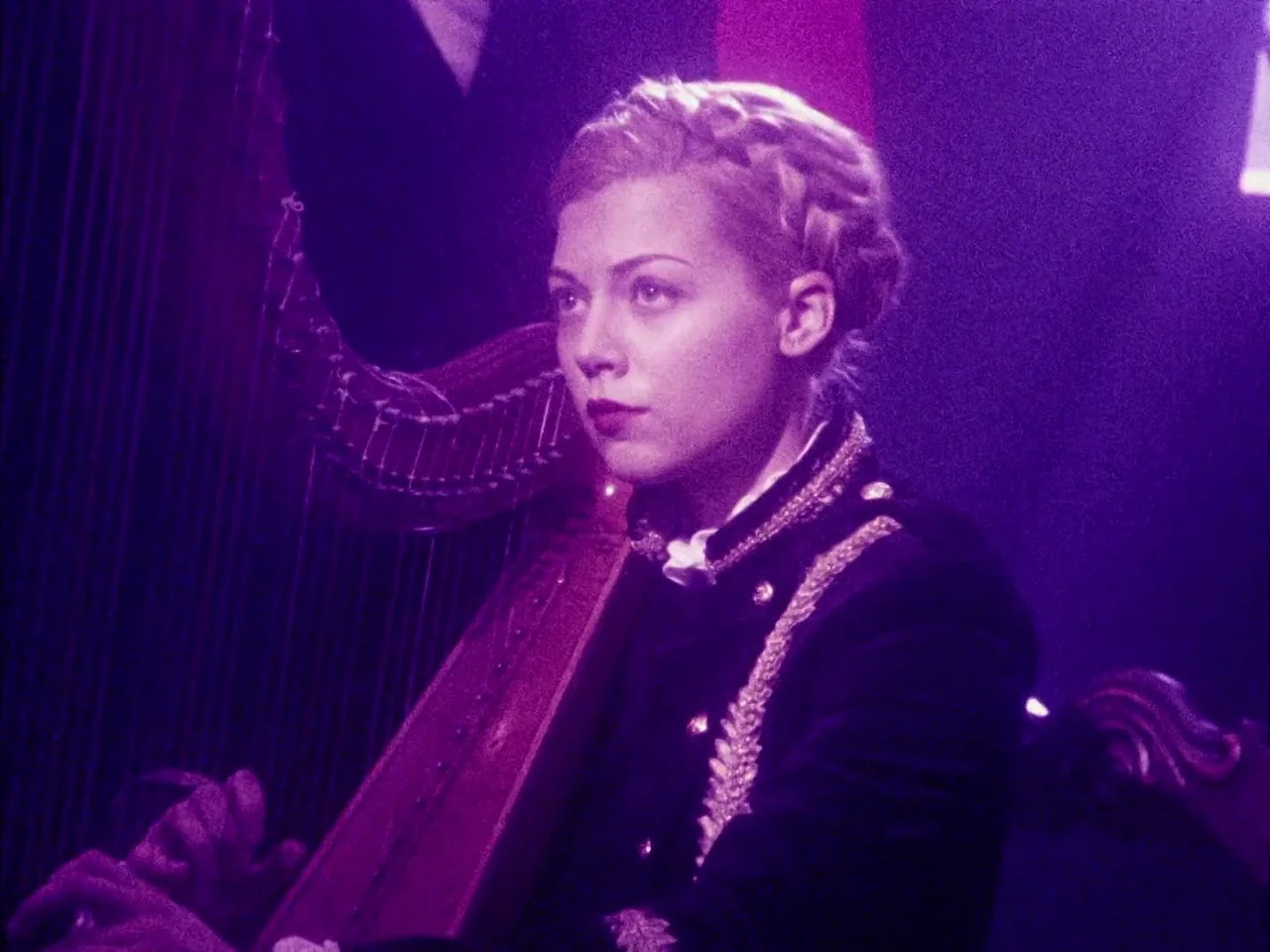 The Twentieth Century (2019) movie still: medium — A woman sits on a stage beside a wooden harp under dim lighting. She wears a braided hairstyle…