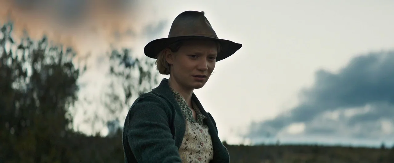 Damsel (2018) movie still: medium — An open rural field under a gray, overcast evening sky. A woman wears a dark coat and hat while looking towards…