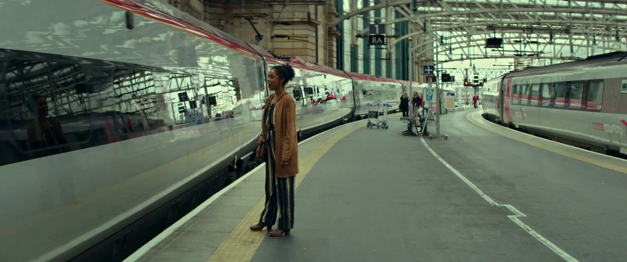Wild Rose (2018) movie still: fullbody — A spacious railway platform features steel train cars parked along both sides of the tracks. A woman…