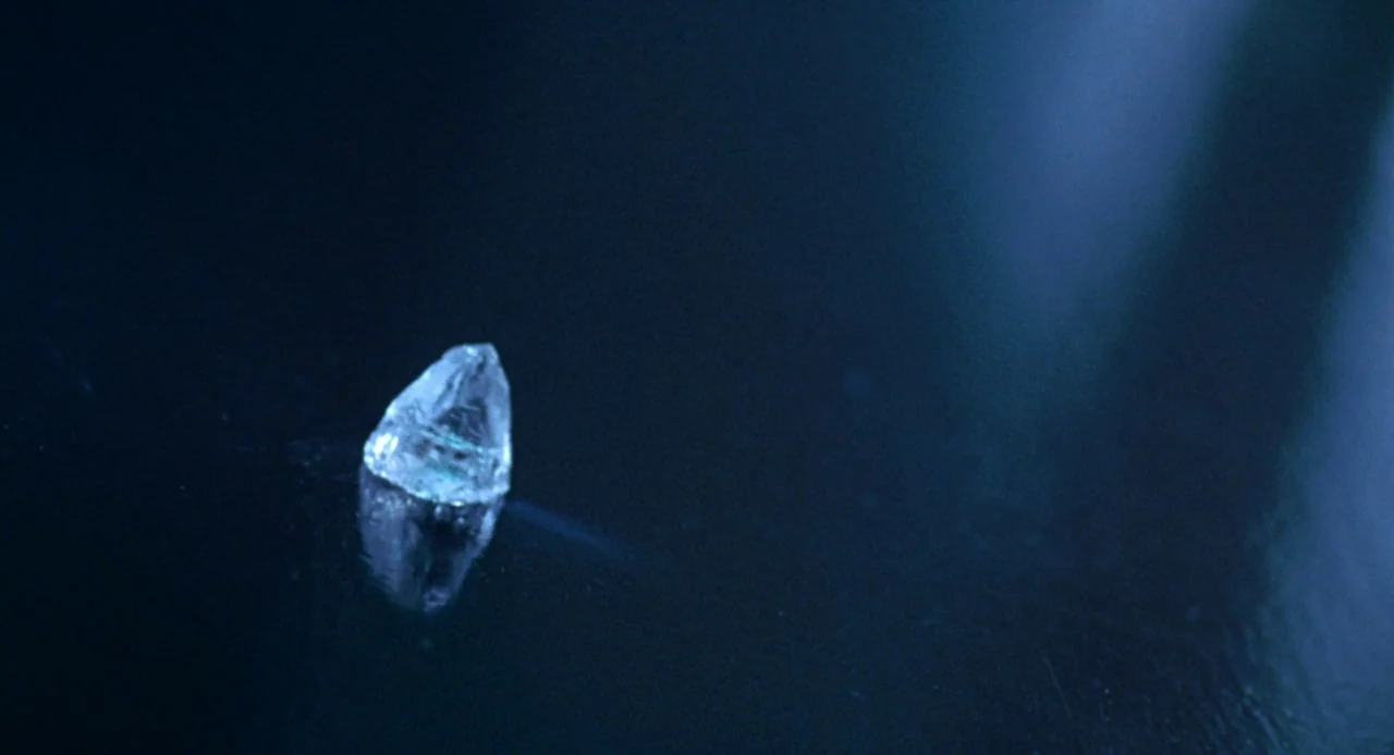 Resident Evil (2002) movie still: closeup — A single clear crystal rests on a dark reflective surface in a dim environment. The stone is illuminated…