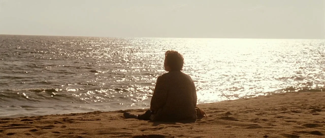 La Vie En Rose (2007) movie still: wide — A sandy beach shoreline borders a vast, sunlit sea under an overcast sky. A person sits on the sand facing…