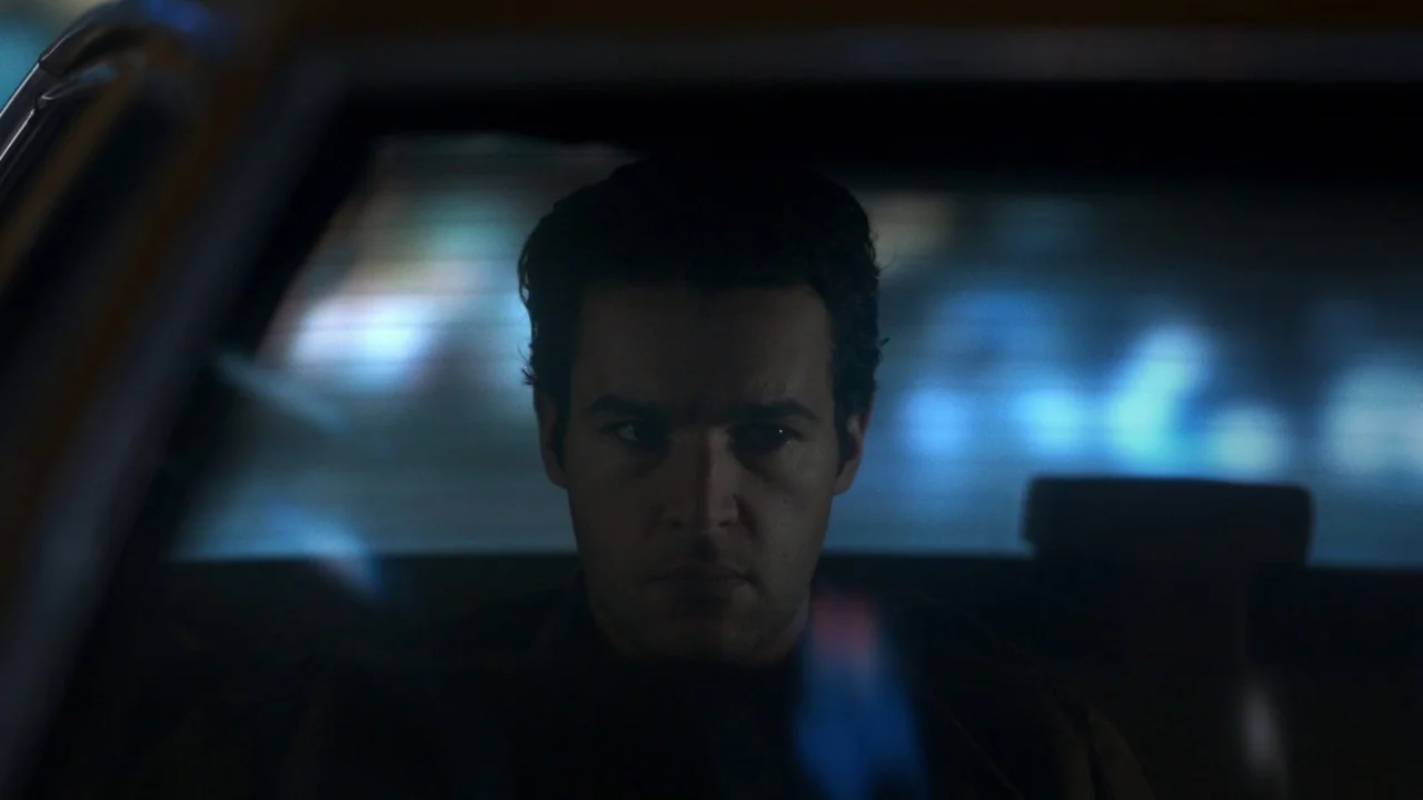 Piercing (2018) movie still: closeup — The interior of a moving car is dark with blue ambient light streaming in. A man with dark hair looks…