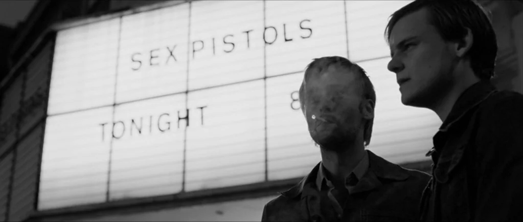 Control movie still: medium — A night exterior features a large illuminated marquee board announcing a Sex Pistols concert. Two men stand in the…
