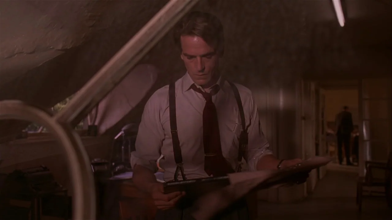 M. Butterfly (1993) movie still: medium — An office space features a sloped ceiling and dim lighting with a desk in view. A man in a dress shirt and…