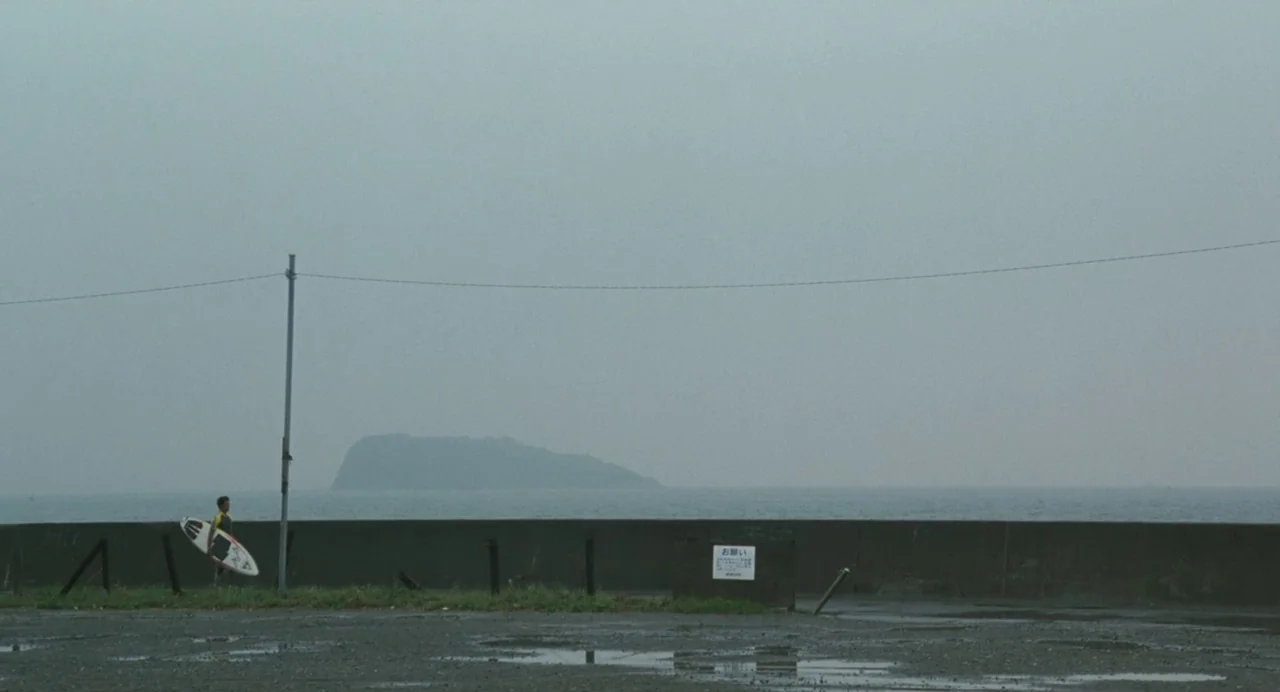 A Scene at the Sea (1991) movie still: wide — An overcast coastal location features a concrete wall separating a parking area from water. A lone…