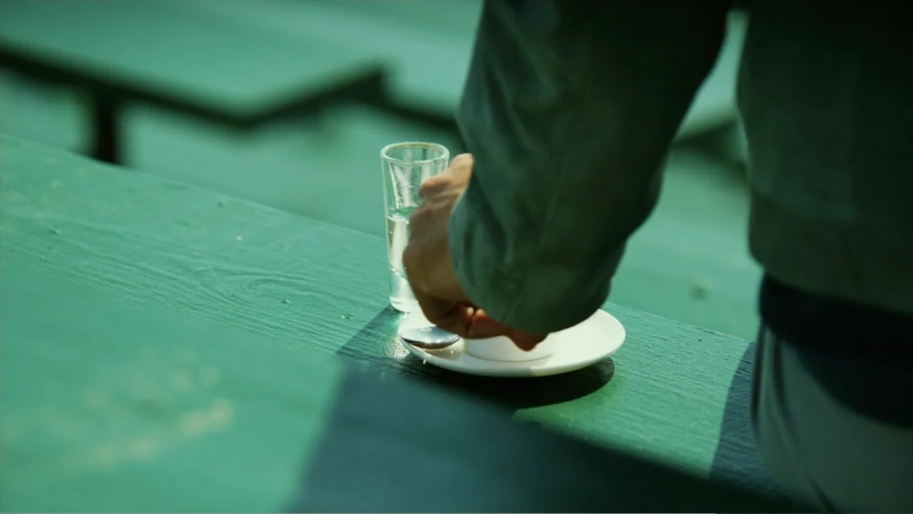 Focus (2015) movie still: medium — A person sits at a green bench at a stadium or outdoor sporting venue. A hand reaches toward a small white cup…