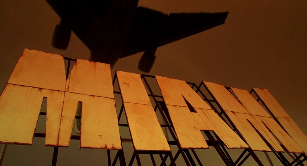 Bad Boys (1995) movie still: establishing — A large sign reading MIAMI sits under a clear, hazy orange sky. A commercial jet aircraft flies low…