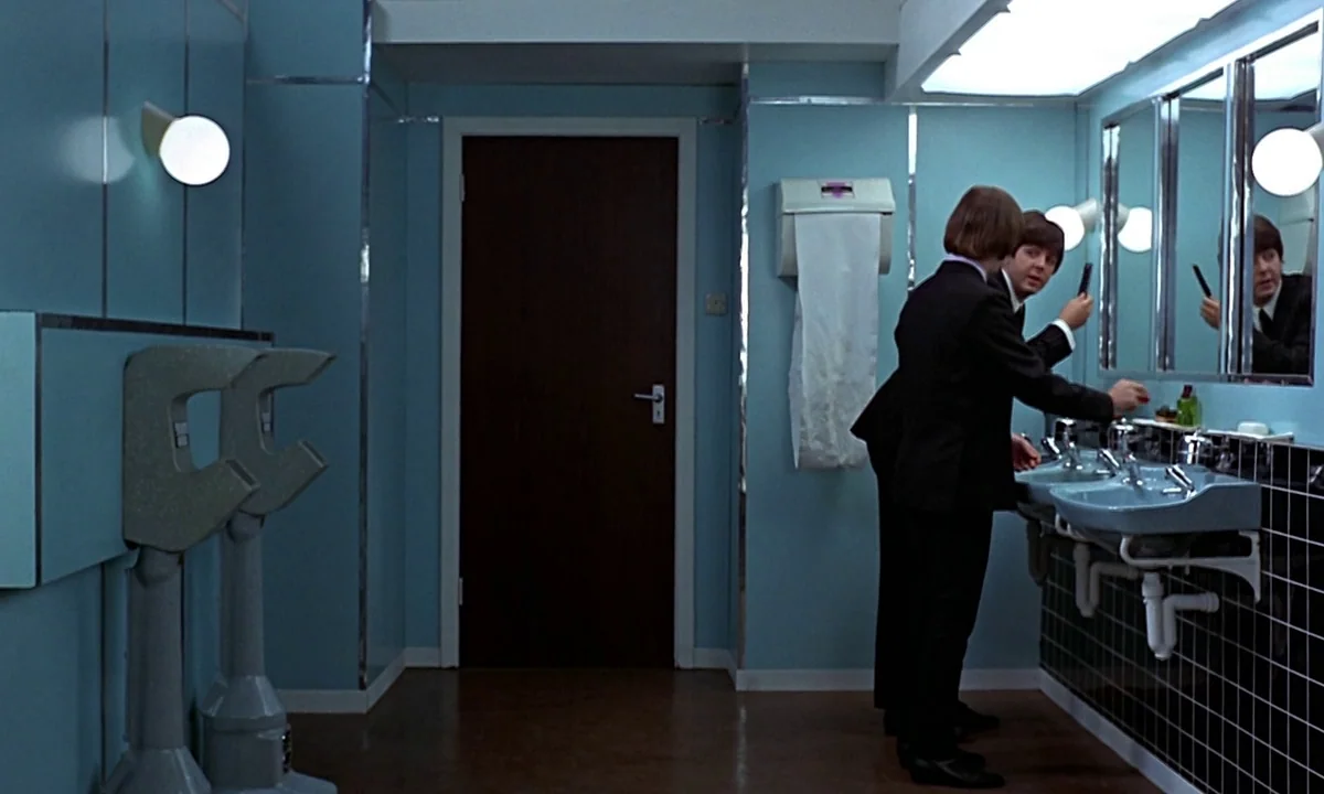Help! (1965) movie still: medium — A 1960s public restroom features light blue walls and a dark wooden door. Two men in dark suits stand before…