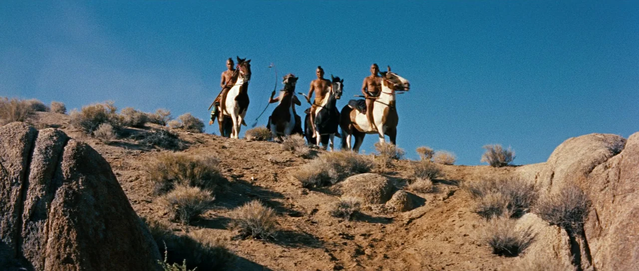 Comanche Station (1960) movie still: wide — A dry, rocky desert slope features sparse scrub brush under a clear blue sky. Three men on horseback…